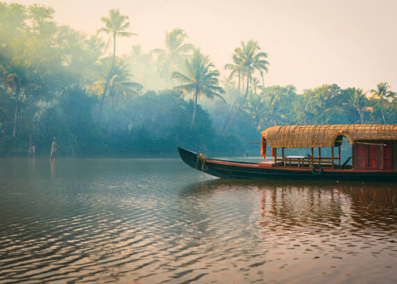A traditional house boat is anchored on the shores of a fishing lake in the palm tree jungle at sunset, in the Backwaters, a popular destination for yoga retreats and nature lovers in Kerala, India